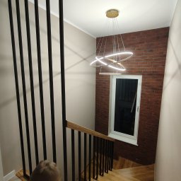HubStal Hubert Czapliński - Nowoczesna balustrada schodowa z czarnymi, pionowymi elementami i drewnianym poręczą, oświetlona designerską lampą wiszącą. Widok z góry na schody.