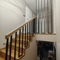 HubStal Hubert Czapliński - Nowoczesne schody z drewnianymi stopniami i czarną, minimalistyczną balustradą. Jasne wnętrze z widocznym oświetleniem LED i fragmentem pomieszczenia poniżej.