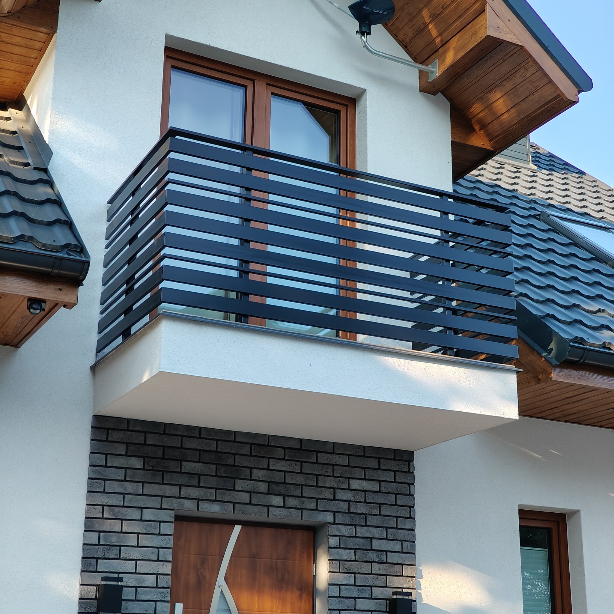 Nowoczesna, pozioma balustrada balkonowa w kolorze antracytowym, zamontowana na białym budynku z elementami dekoracyjnymi z cegły i drewnianymi okładzinami dachu.