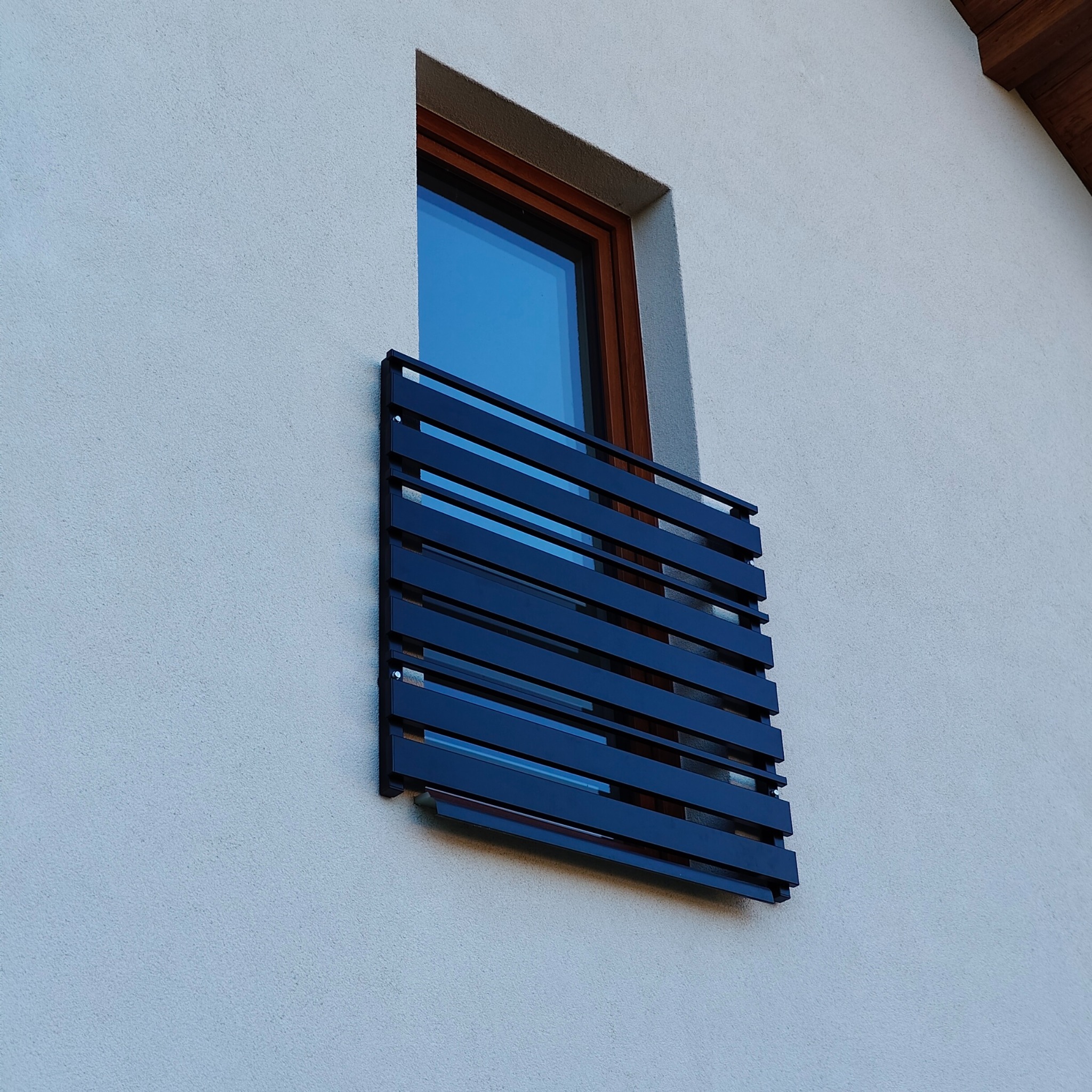 Nowoczesna, pozioma balustrada balkonowa w kolorze antracytowym, zamontowana przy oknie na elewacji budynku. Minimalistyczny design, widok z dołu.