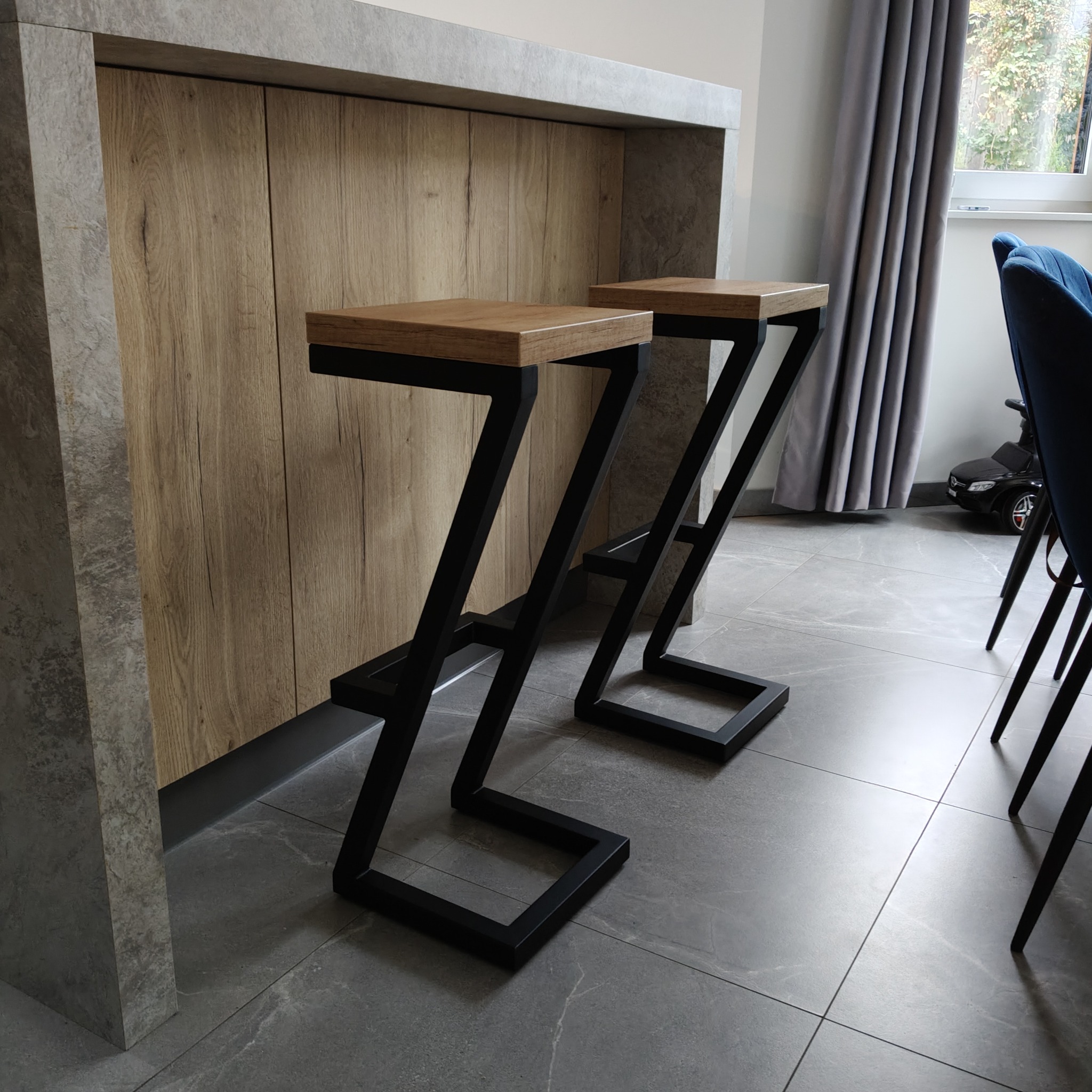 Dwa nowoczesne hokery barowe z drewnianymi siedziskami i czarnymi, metalowymi nogami w kształcie litery Z, ustawione przy blacie kuchennym z drewnianymi frontami.