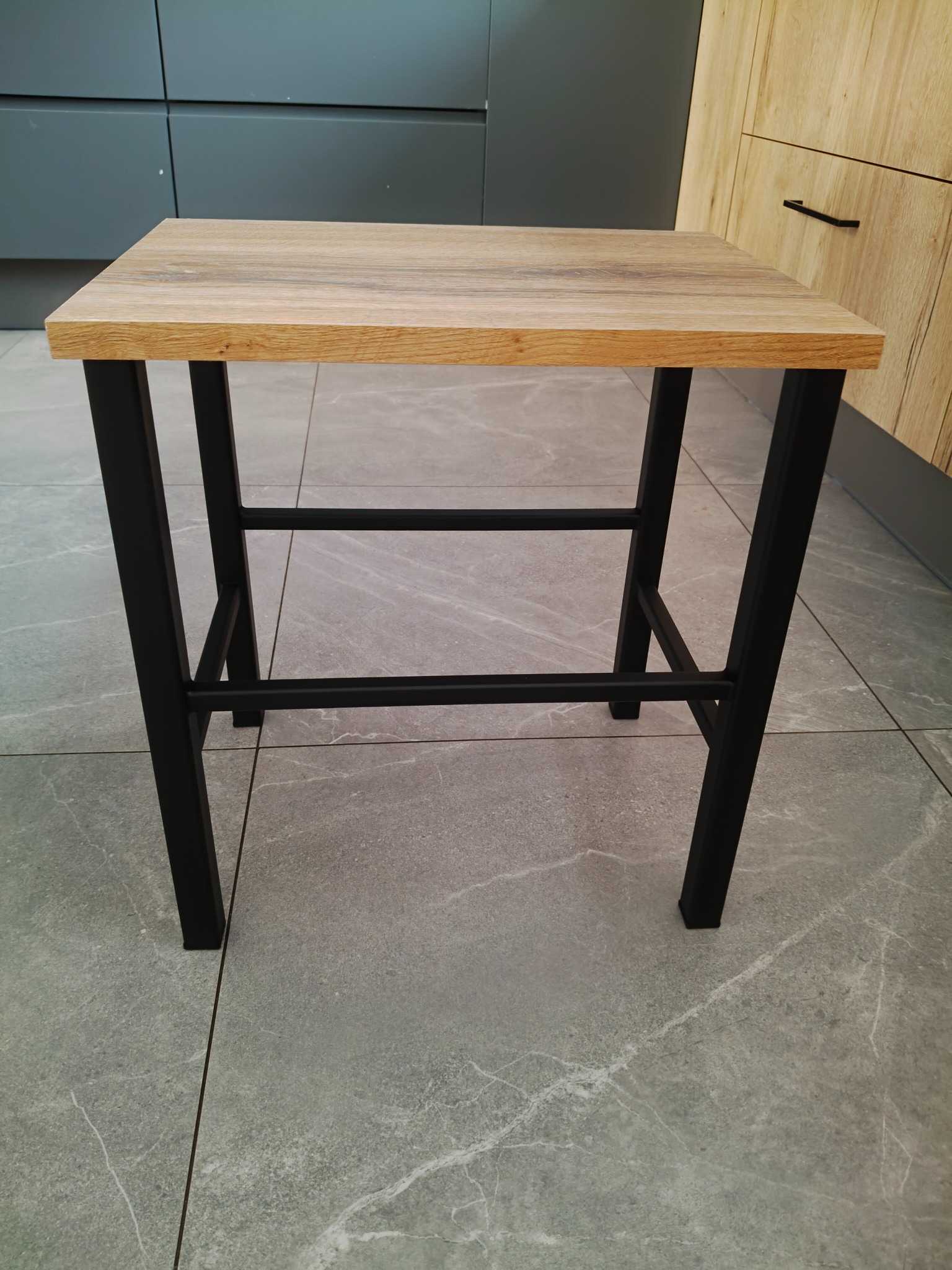 Stolik kawowy z blatem imitującym drewno i czarnymi, metalowymi nogami, na tle szarej podłogi i zabudowy kuchennej w odcieniach szarości i drewna.