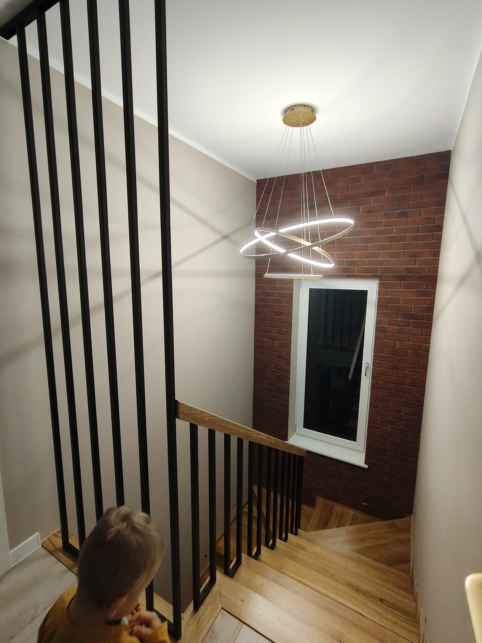 Nowoczesna balustrada schodowa z czarnymi, pionowymi elementami i drewnianym poręczą, oświetlona designerską lampą wiszącą. Widok z góry na schody.