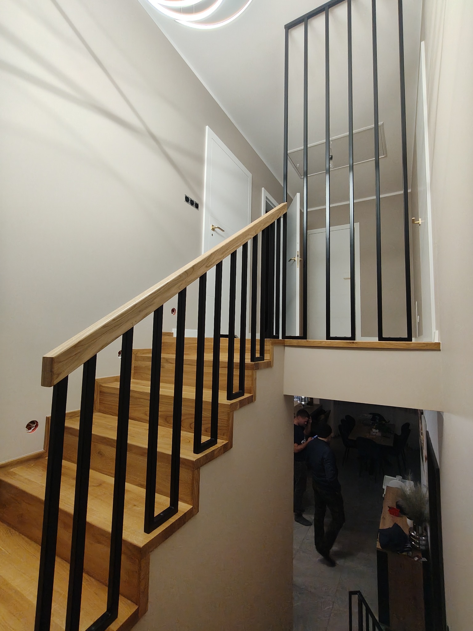 Nowoczesne schody z drewnianymi stopniami i czarną, minimalistyczną balustradą. Jasne wnętrze z widocznym oświetleniem LED i fragmentem pomieszczenia poniżej.