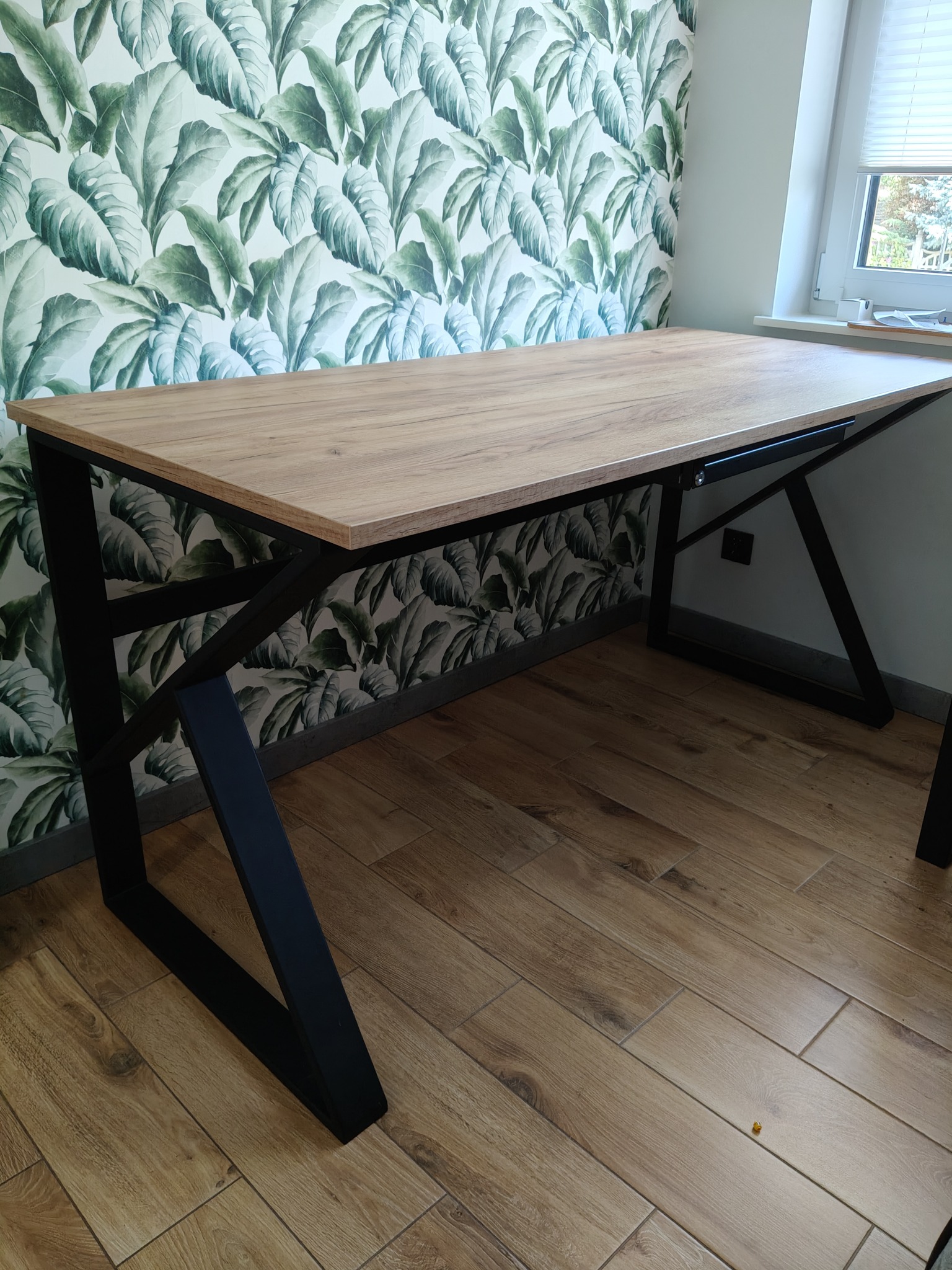 Stół z blatem imitującym drewno i czarnymi, metalowymi nogami o geometrycznym kształcie na tle ściany w liście. Widoczne płytki podłogowe imitujące drewno.