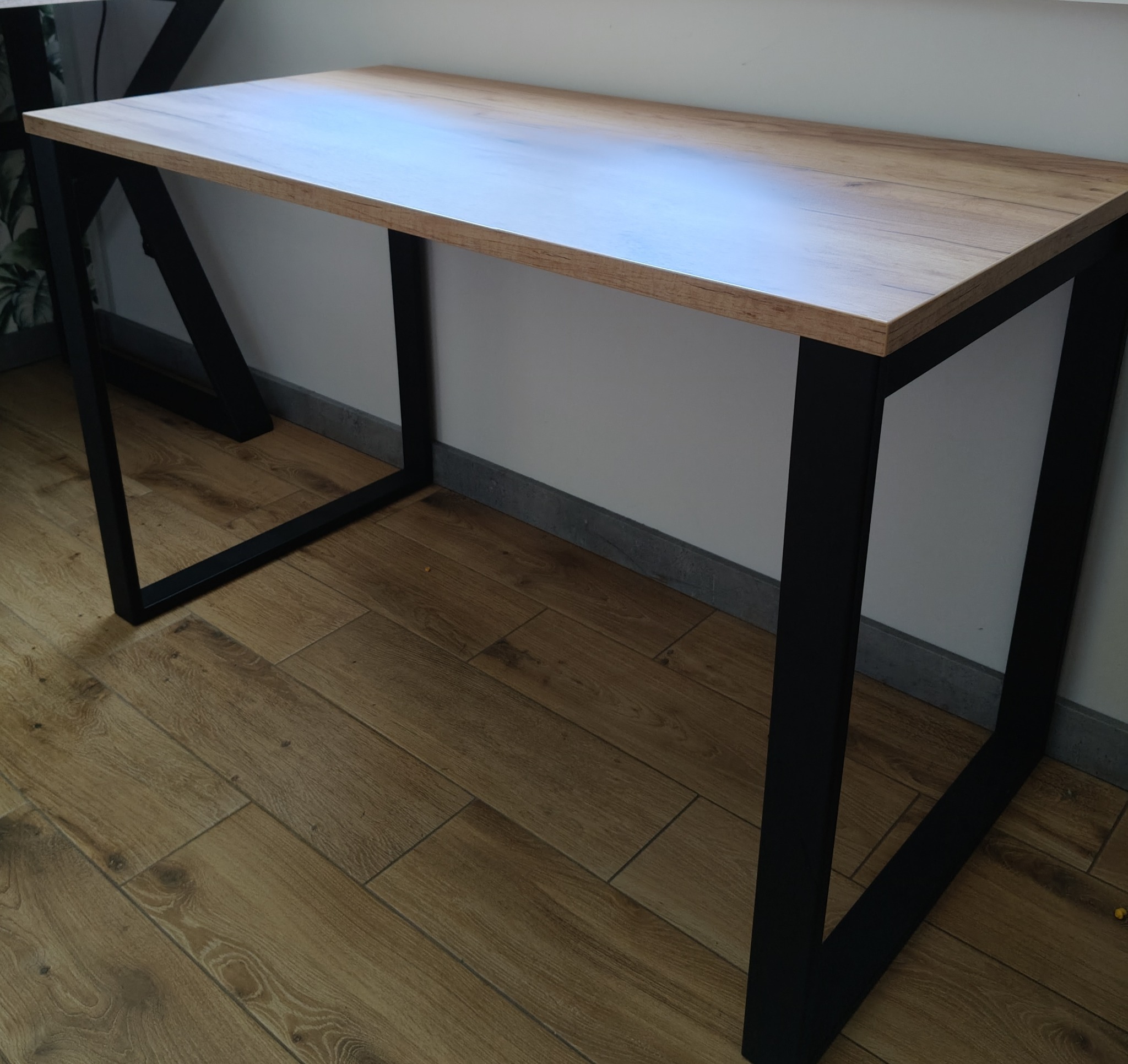 Biurko z blatem imitującym drewno i czarnymi, metalowymi nogami o geometrycznym kształcie, stojące na podłodze z paneli. Minimalistyczny design.
