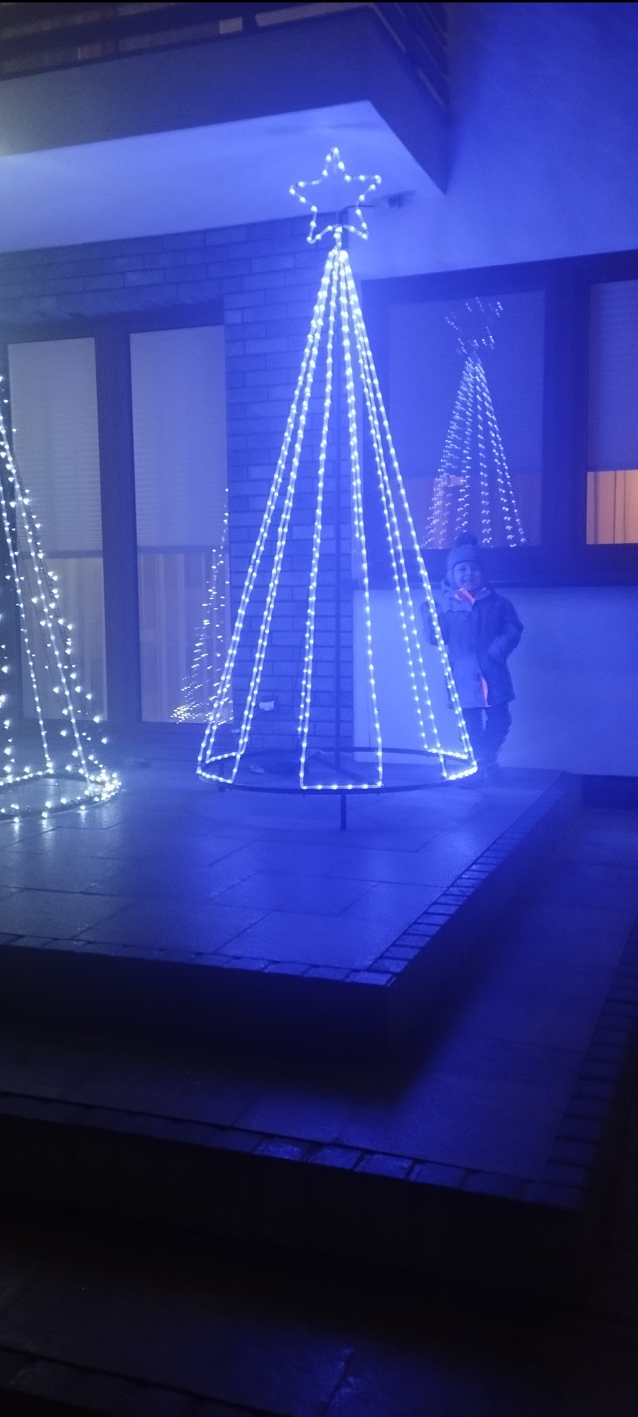 Iluminowana, metalowa konstrukcja choinki z gwiazdą na szczycie, oświetlona niebieskim światłem, z dzieckiem w tle, odbijającym się w oknie.