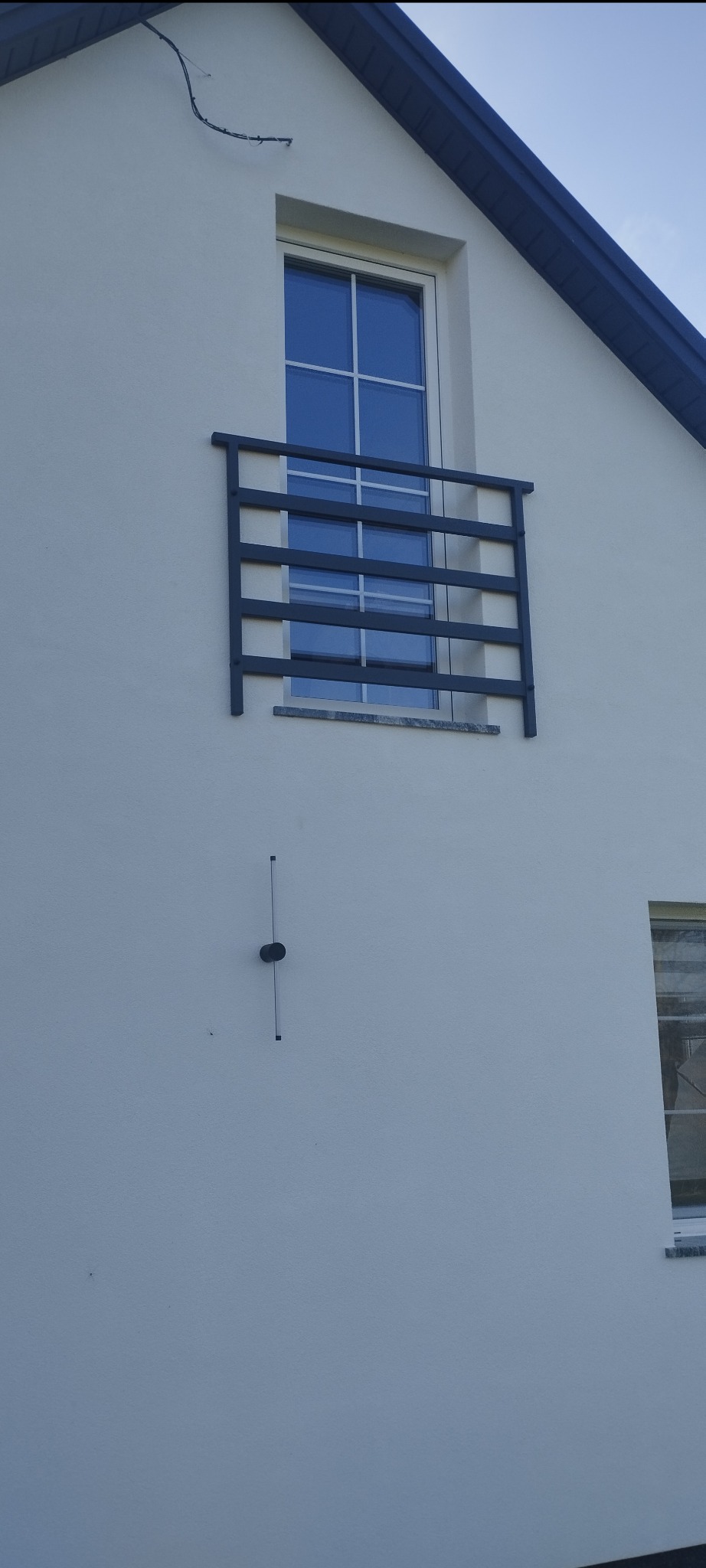 Szara, metalowa balustrada okienna na białej elewacji domu z oknem. Minimalistyczny design, proste linie. Widok z dołu, jasne tło nieba.