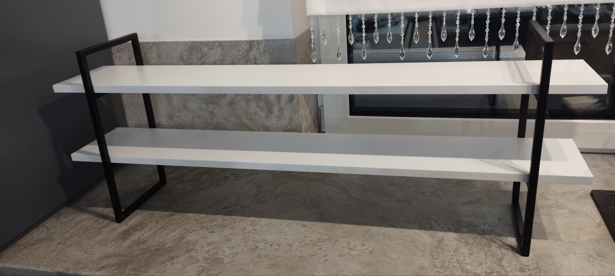 Nowoczesny regał z dwoma białymi półkami i czarną metalową ramą, ustawiony na blacie w pomieszczeniu z oknem w tle, zasłoniętym dekoracyjną zasłoną.