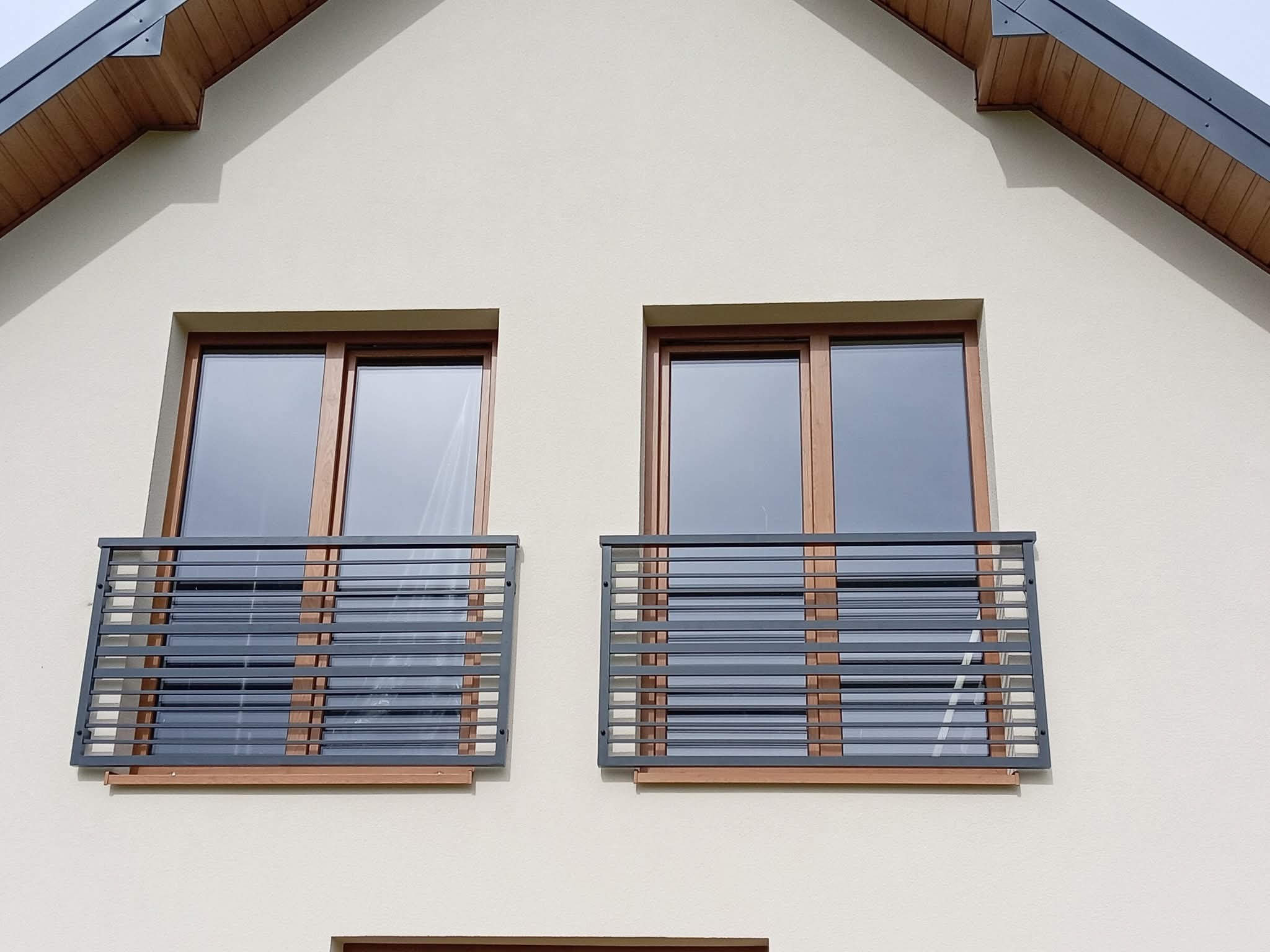 Dwa balkony francuskie z szarą, poziomą balustradą na elewacji domu. Drewniane ramy okienne i podbitka dachu dodają ciepła. Minimalistyczny design.