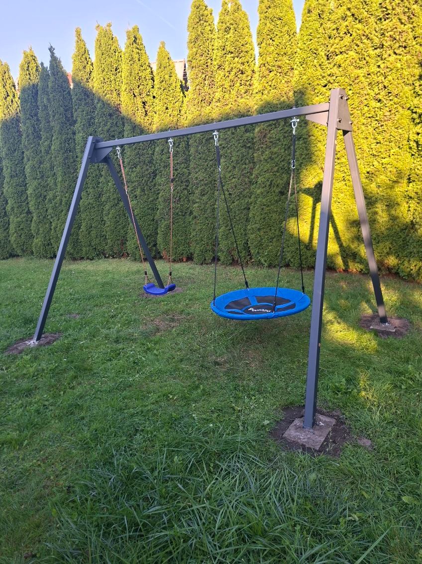 Metalowa huśtawka ogrodowa z dwoma siedziskami: okrągłym niebieskim i klasycznym. W tle wysokie tuje i trawnik. Konstrukcja pomalowana na szaro.
