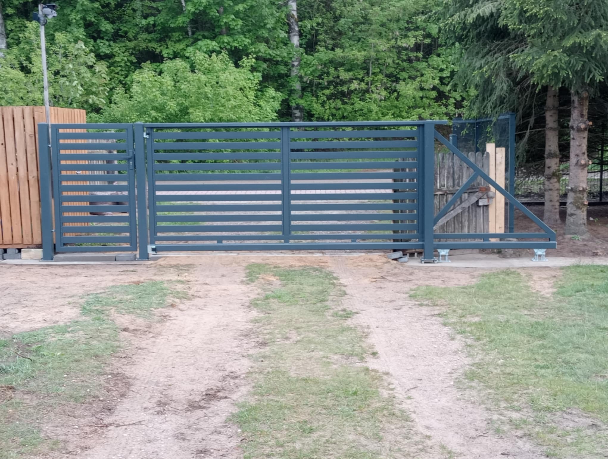 Nowoczesna brama wjazdowa z poziomymi panelami, pomalowana na szaro, z widocznym mechanizmem przesuwnym. Tło stanowi zielony las i fragment drewnianego ogrodzenia.