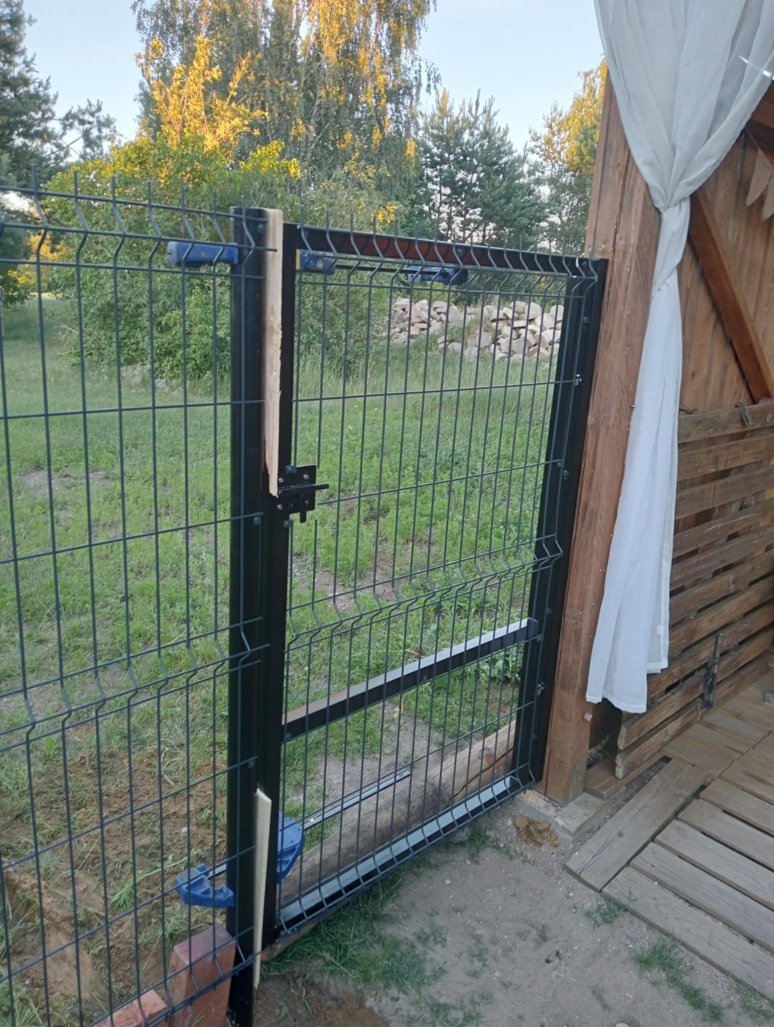 Nowoczesna furtka z metalowej siatki w Bargłowie Kościelnym, z widocznym zamkiem i elementami mocującymi, prowadząca do ogrodu z drewnianą konstrukcją w tle.