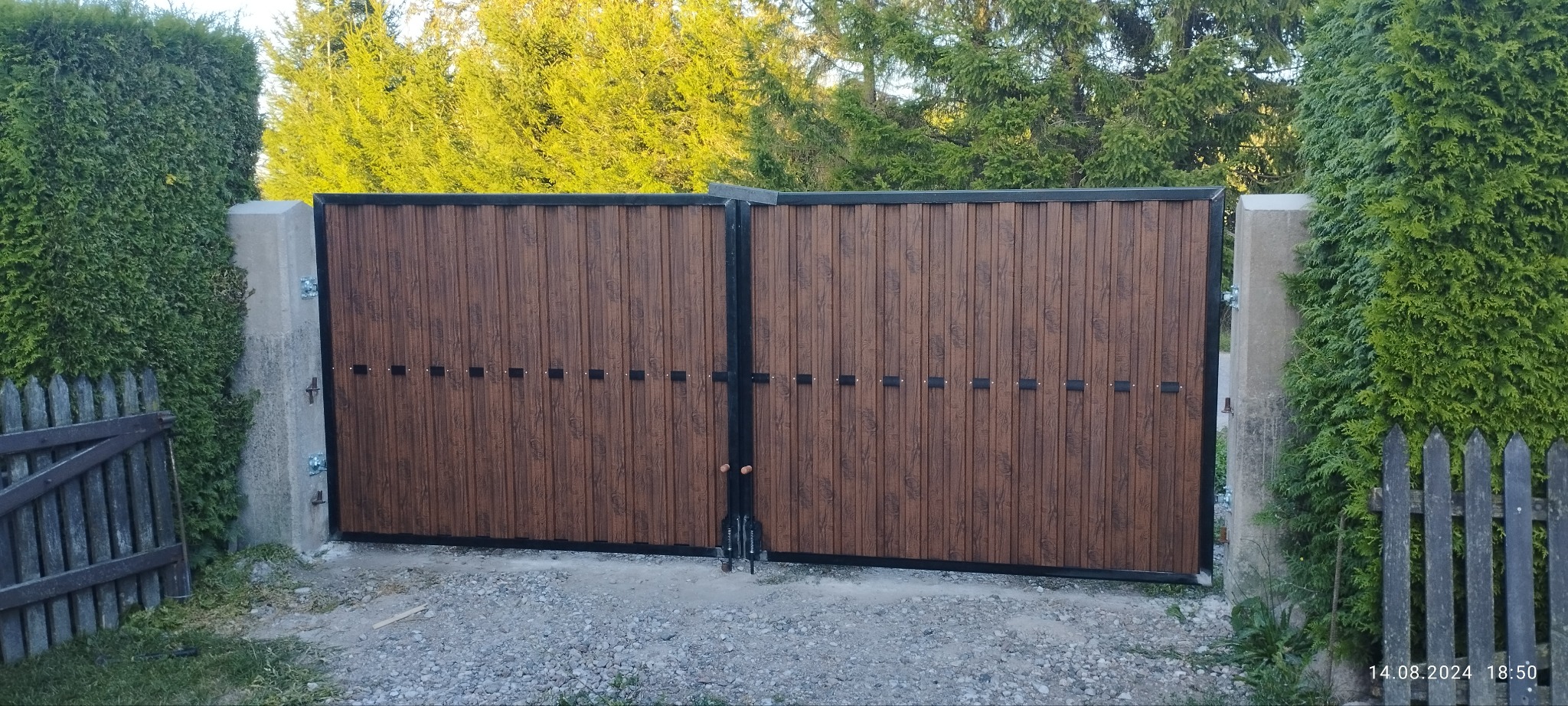 Dwuskrzydłowa brama wjazdowa z brązowymi panelami pionowymi i czarną ramą, osadzona między betonowymi słupkami, otoczona zielenią.