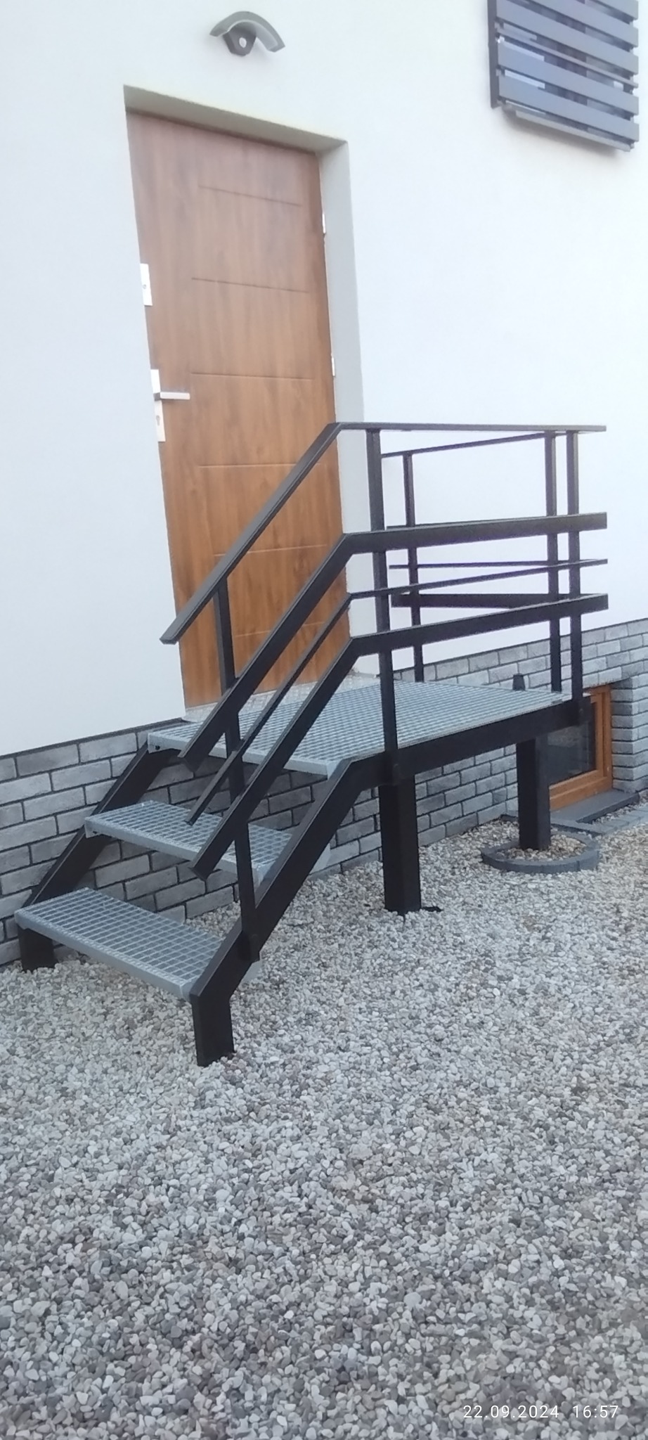Metalowe schody zewnętrzne z balustradą, prowadzące do drzwi wejściowych. Schody z kraty pomostowej, czarna konstrukcja, jasne kamienie na podłożu.