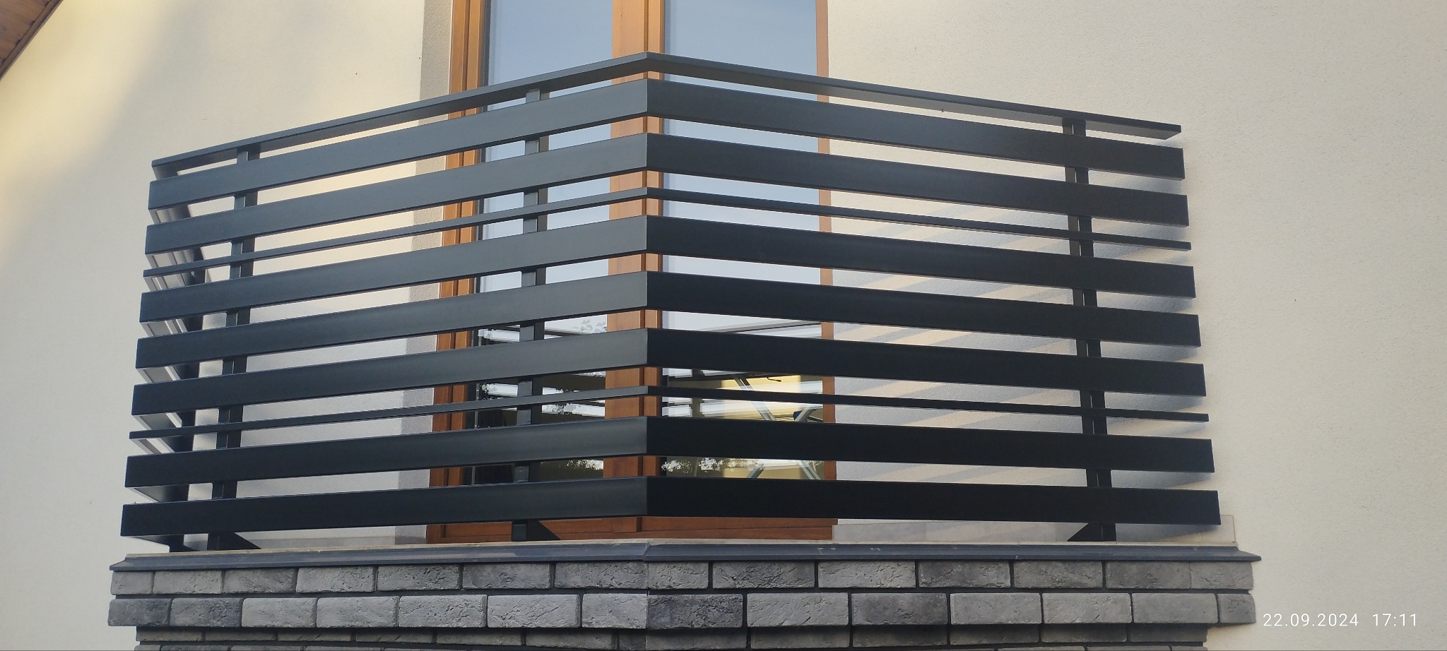 Nowoczesna, czarna balustrada balkonowa z poziomych profili, zamontowana na murku z szarej cegły. Widoczne okno w tle. Minimalistyczny design.