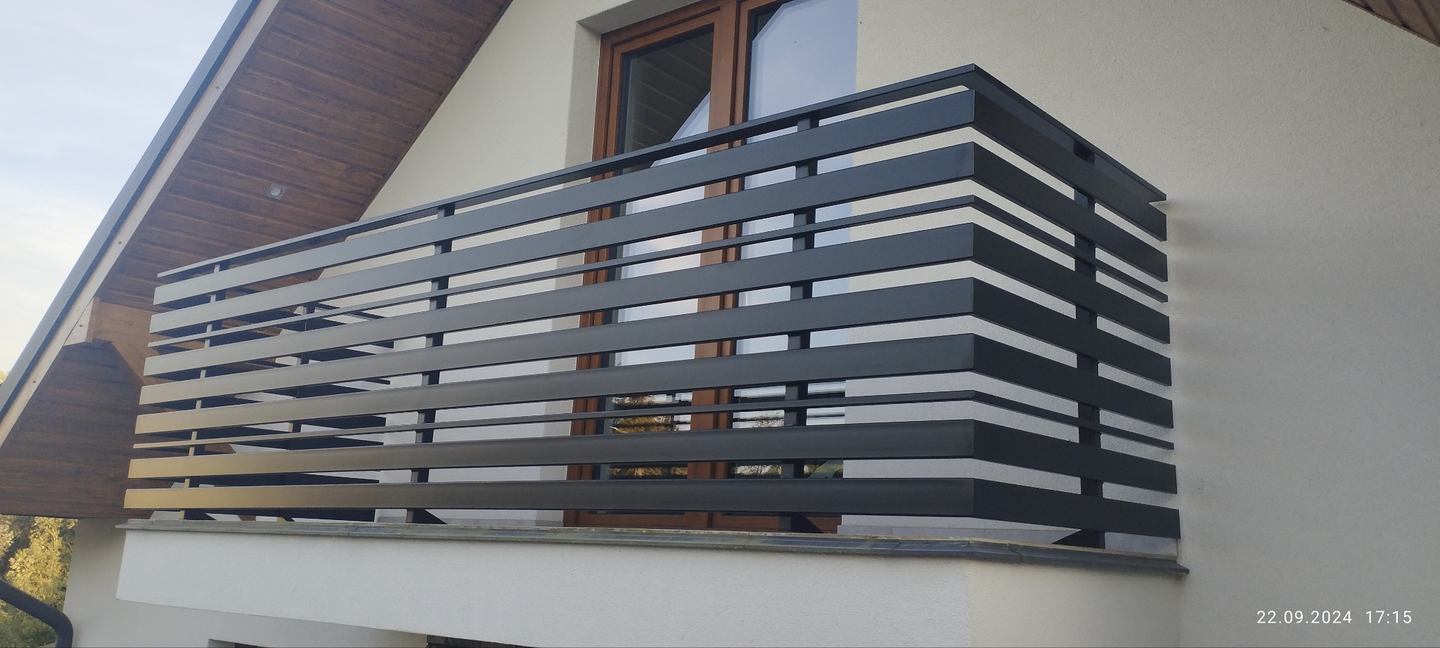 Nowoczesna, pozioma balustrada balkonowa w kolorze antracytowym, z widocznym fragmentem elewacji domu i drewnianym wykończeniem dachu. Minimalistyczny design.