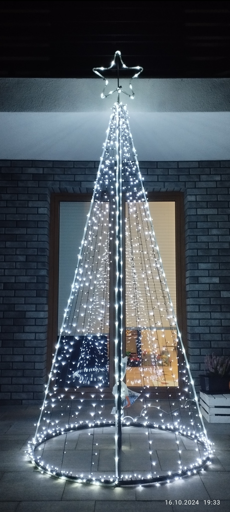Metalowa konstrukcja w kształcie choinki, opleciona białymi światełkami LED, zwieńczona gwiazdą. Stoi na zewnątrz, na tle ceglanego muru i okna.