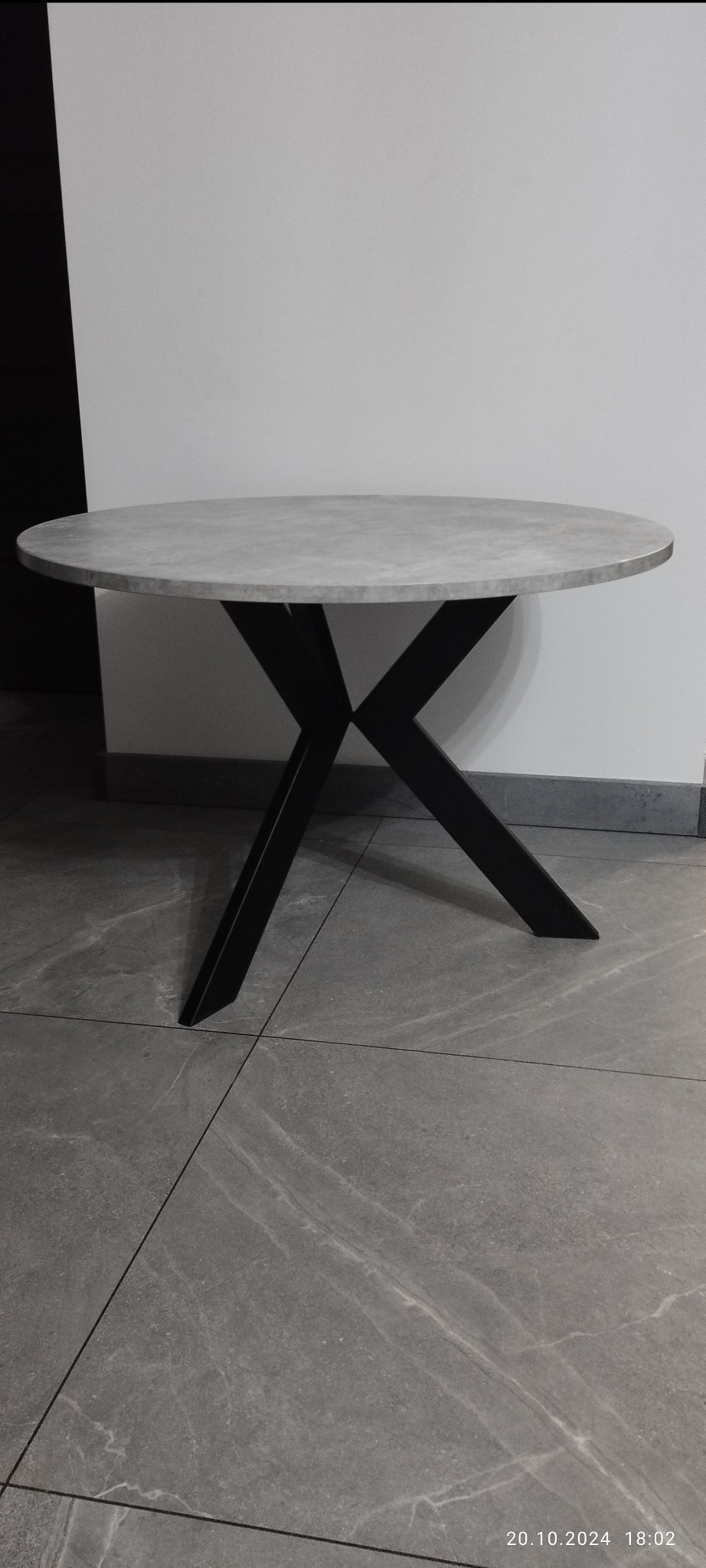 Stół z okrągłym blatem imitującym beton i czarną, metalową podstawą w kształcie litery X, na tle białej ściany i szarej podłogi z płytek.