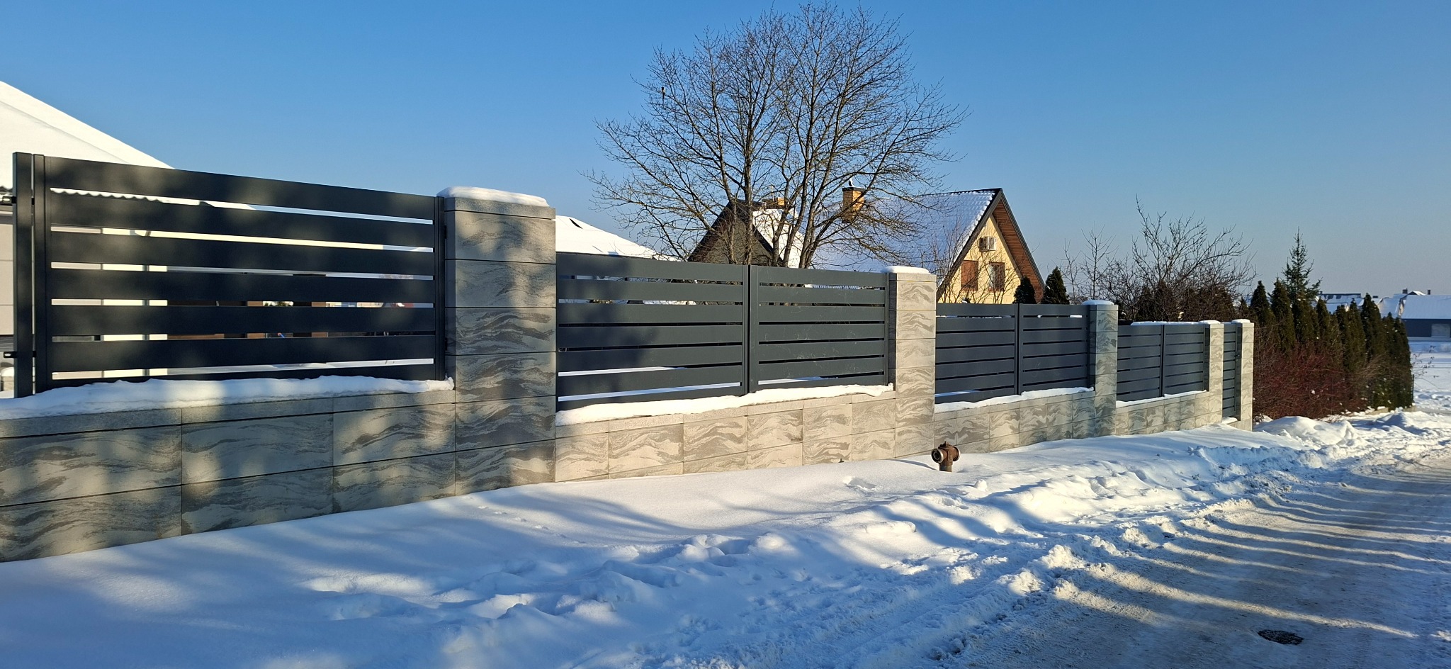 Nowoczesne ogrodzenie panelowe z szarego metalu i kamiennych słupków w zimowej scenerii. Widoczne zaspy śnieżne i dom w tle. Słoneczny dzień.