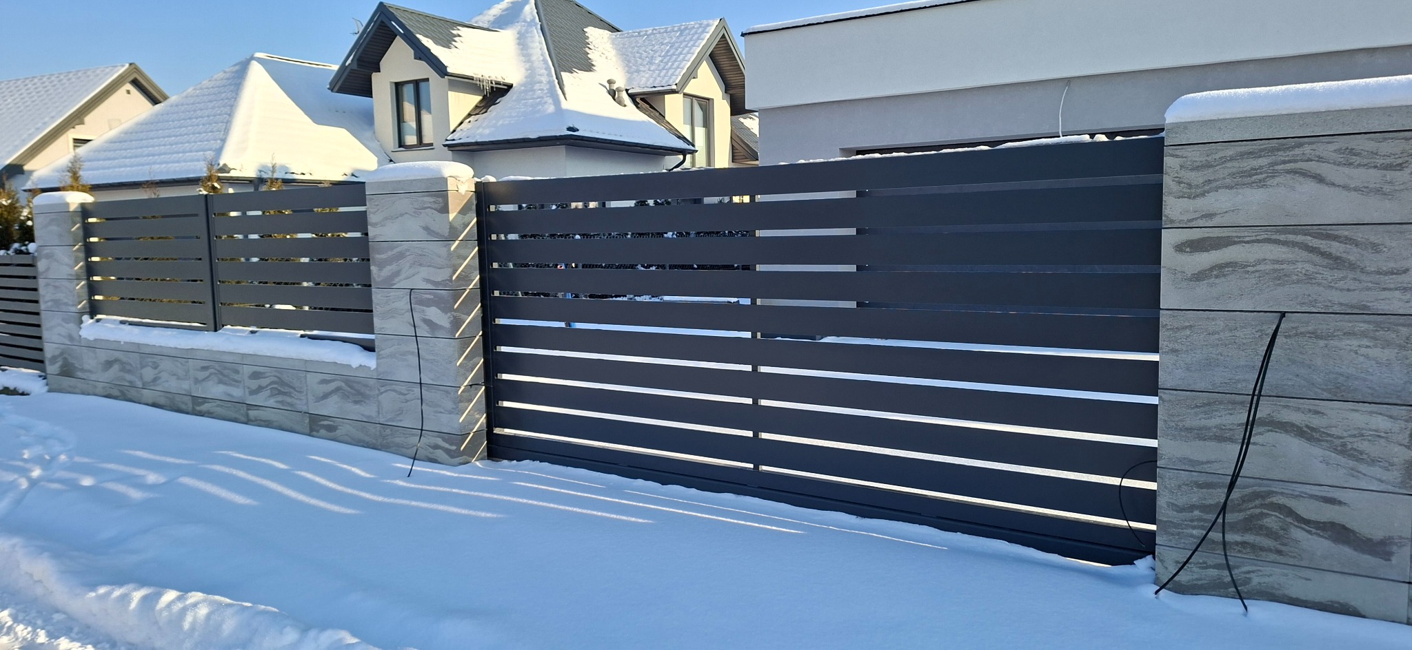 Nowoczesna, pozioma brama wjazdowa w kolorze antracytu z kamiennymi słupkami, pokryta śniegiem. W tle domy jednorodzinne pod błękitnym niebem.