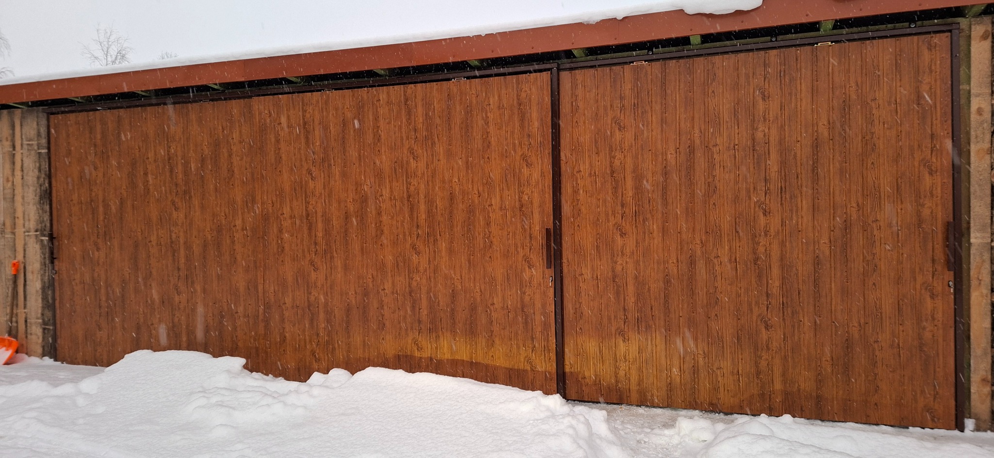 Drewniana brama przesuwna w zimowej scenerii. Widoczne deski z wyraźnym usłojeniem i metalowa rama. Śnieg leży u podstawy bramy, a na górze widać fragment dachu.