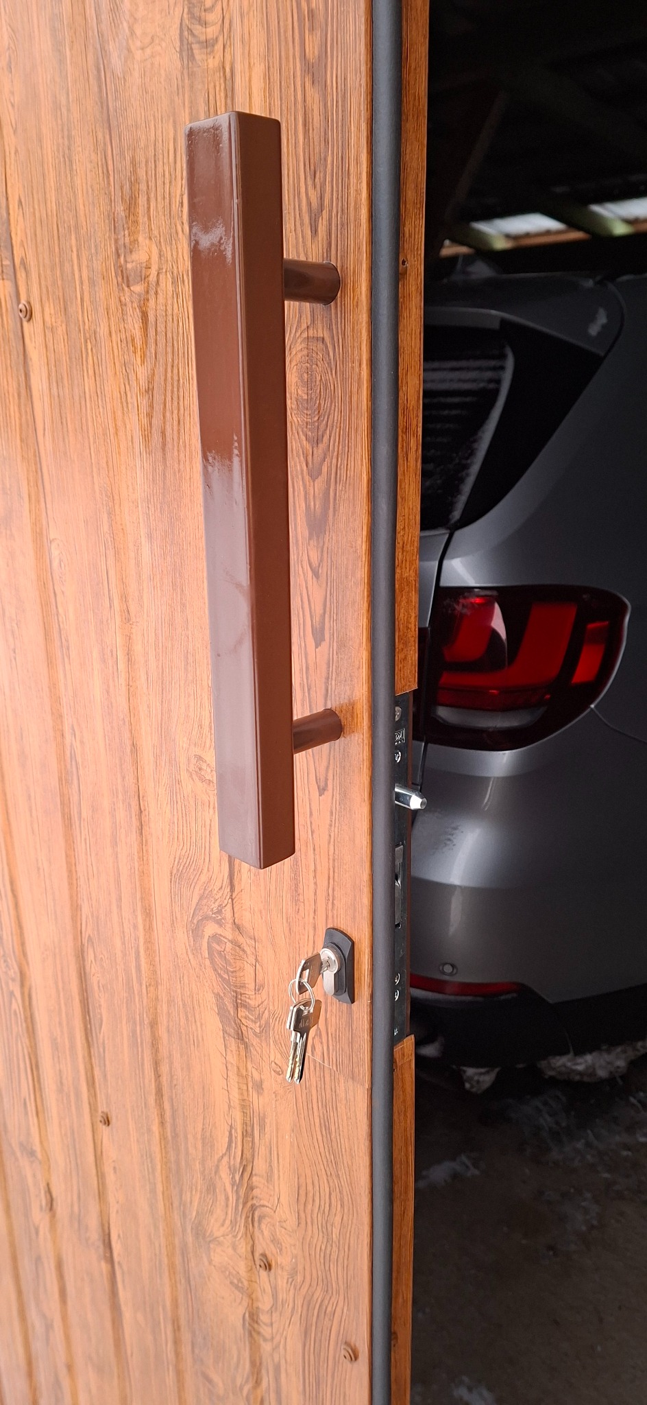 Otwarta, brązowa brama z drewnopodobnym wzorem i dużą, prostokątną klamką. W zamku tkwi klucz. W tle widoczny fragment samochodu.