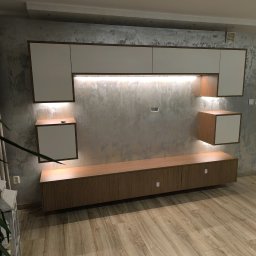 Komfortis Meble Tomasz Kmieć - Nowoczesna zabudowa meblowa w salonie z oświetleniem LED. Białe szafki wiszące z drewnianymi akcentami na tle ściany o fakturze betonu. Drewniana podłoga.