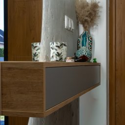 Komfortis Meble Tomasz Kmieć - Półka wisząca z szufladą w odcieniach drewna i szarości, dekorowana wazonami i świecami, na tle ściany o fakturze betonu i drewnianych drzwi.