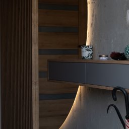 Komfortis Meble Tomasz Kmieć - Nowoczesna szafa wnękowa z oświetleniem LED i drewnianymi elementami dekoracyjnymi. Na półce ozdobne przedmioty i parasol. Betonowa ściana w tle.