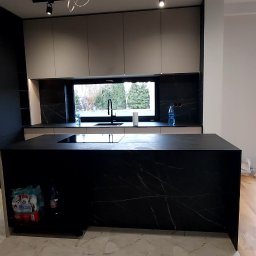 Komfortis Meble Tomasz Kmieć - Nowoczesna kuchnia z czarną wyspą i marmurowym blatem, widok na okno. Minimalistyczny design, oświetlenie punktowe, butelki wody pod blatem.