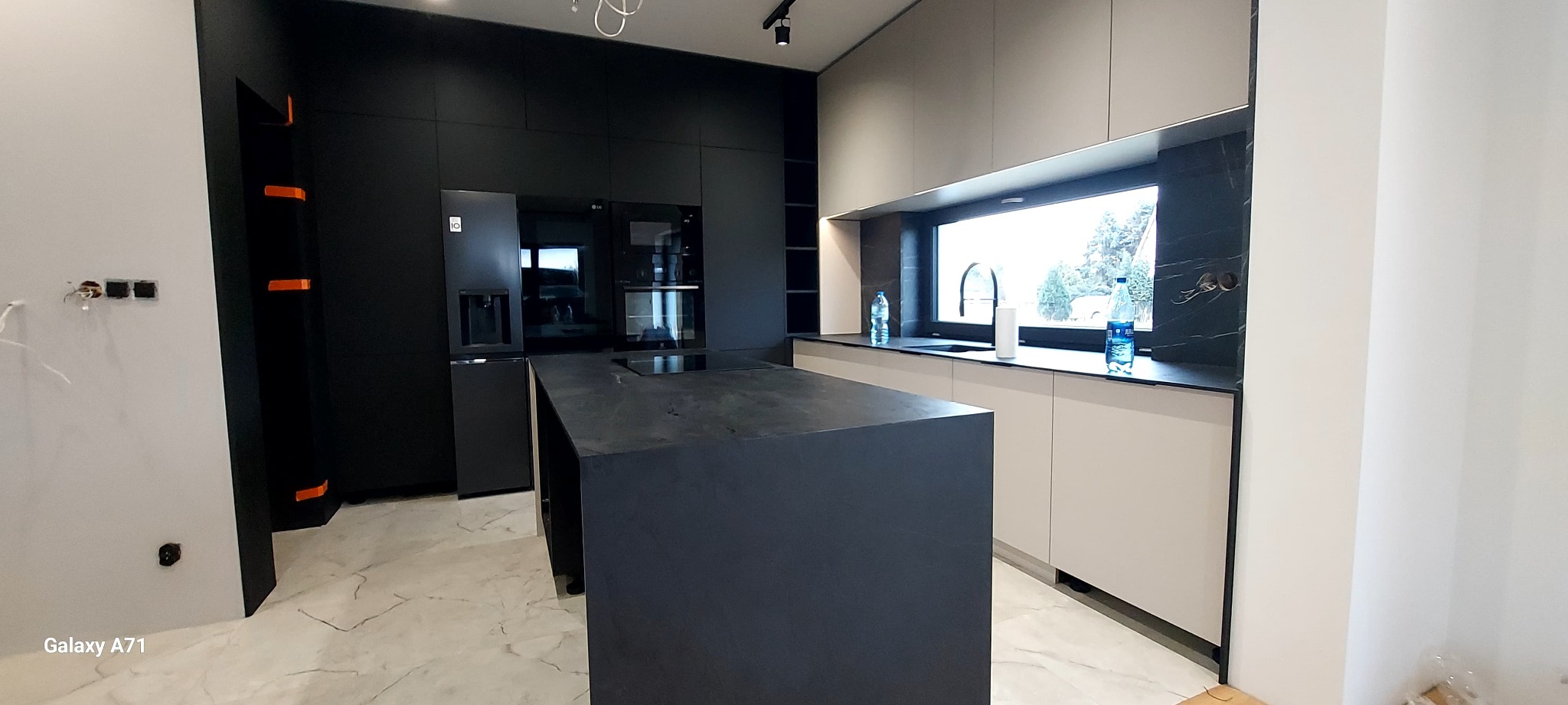 Nowoczesna kuchnia z wyspą: czarne fronty szafek, beżowe dolne szafki, marmurowy blat i okno z widokiem na zieleń. Minimalistyczny design, oświetlenie punktowe.