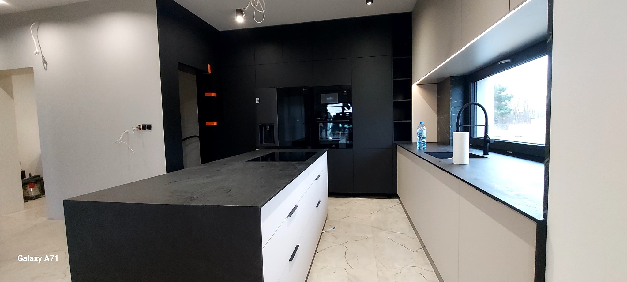 Nowoczesna kuchnia z wyspą: czarne szafki górne, białe dolne, marmurowa podłoga. Minimalistyczny design, czarne akcenty, okno z widokiem na zewnątrz.