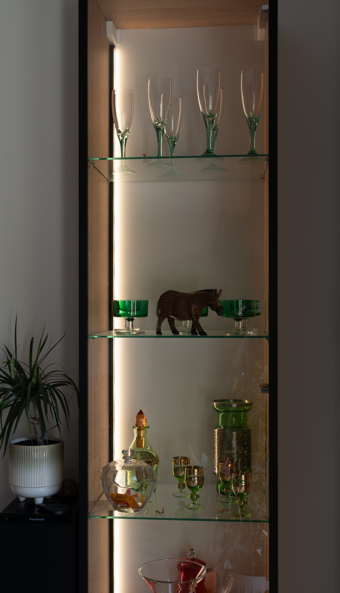 Podświetlana szafa wnękowa z przeszklonymi półkami, na których stoją dekoracyjne kieliszki, wazony i figurka zebry. Obok szafy widoczny kwiat doniczkowy.