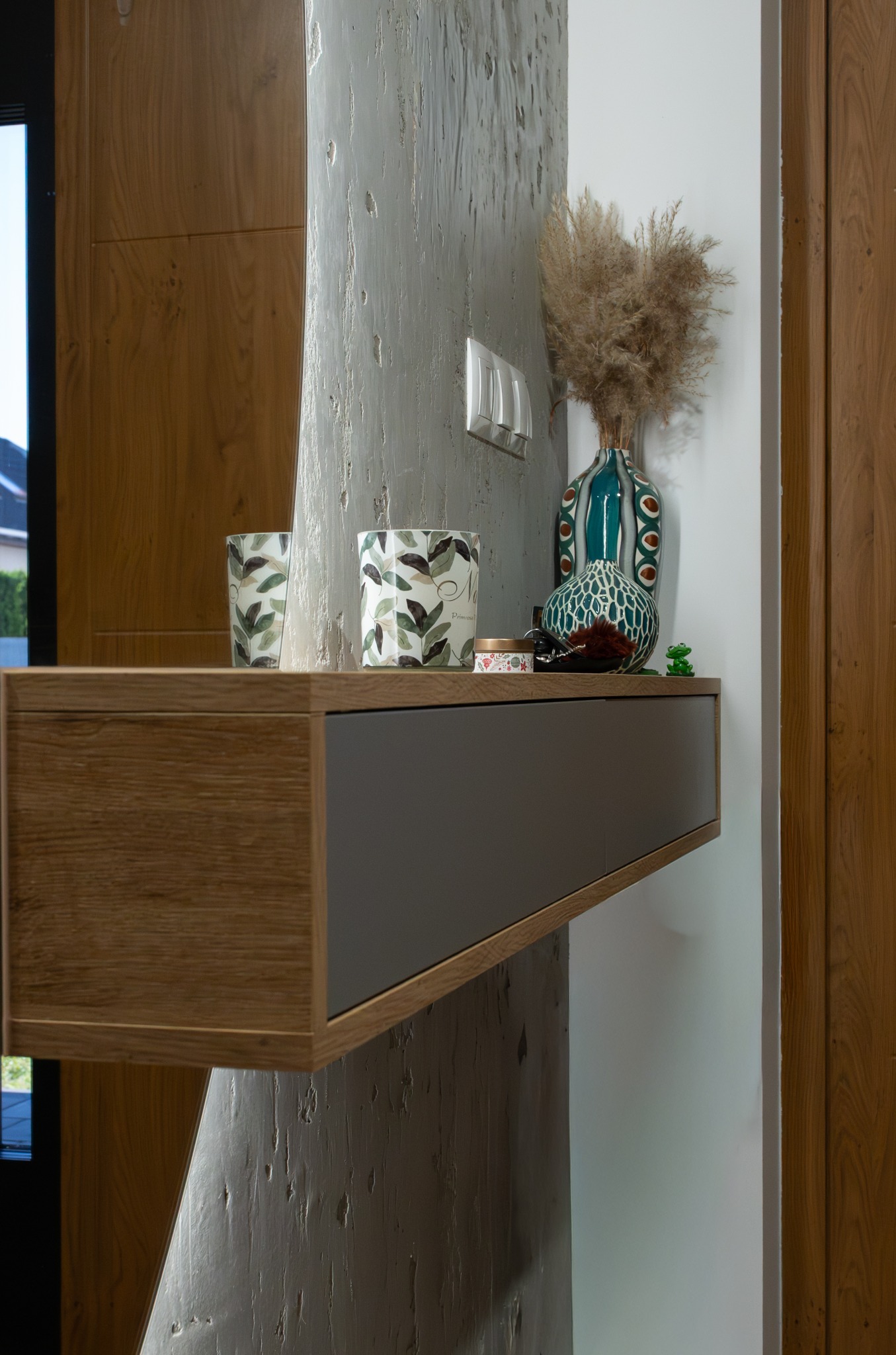 Półka wisząca z szufladą w odcieniach drewna i szarości, dekorowana wazonami i świecami, na tle ściany o fakturze betonu i drewnianych drzwi.