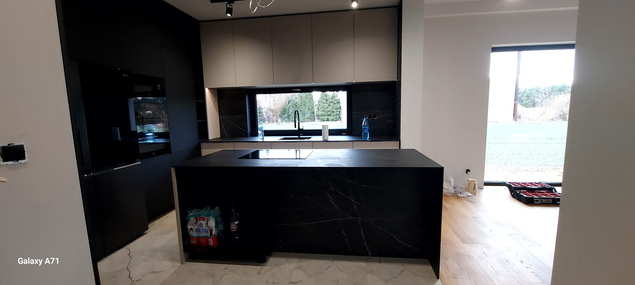 Nowoczesna kuchnia z czarną wyspą i marmurowym blatem, widok na okno. Minimalistyczny design, oświetlenie punktowe, butelki wody pod blatem.
