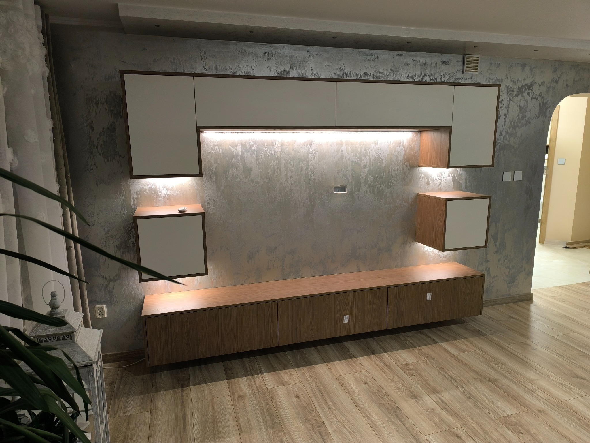 Nowoczesna szafka RTV z białymi frontami i drewnianymi elementami, podświetlona listwą LED, na tle ściany z efektem betonu i drewnianej podłogi.