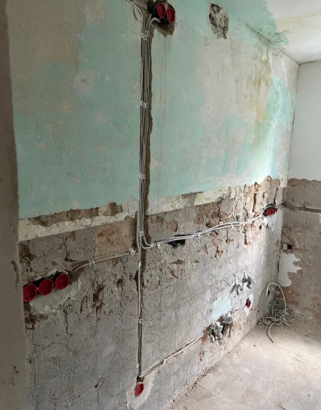 Ściana w trakcie remontu z widoczną instalacją elektryczną: kable w peszlach i czerwone puszki pod gniazdka. Częściowo skuwa ściana, widoczne cegły i resztki farby.