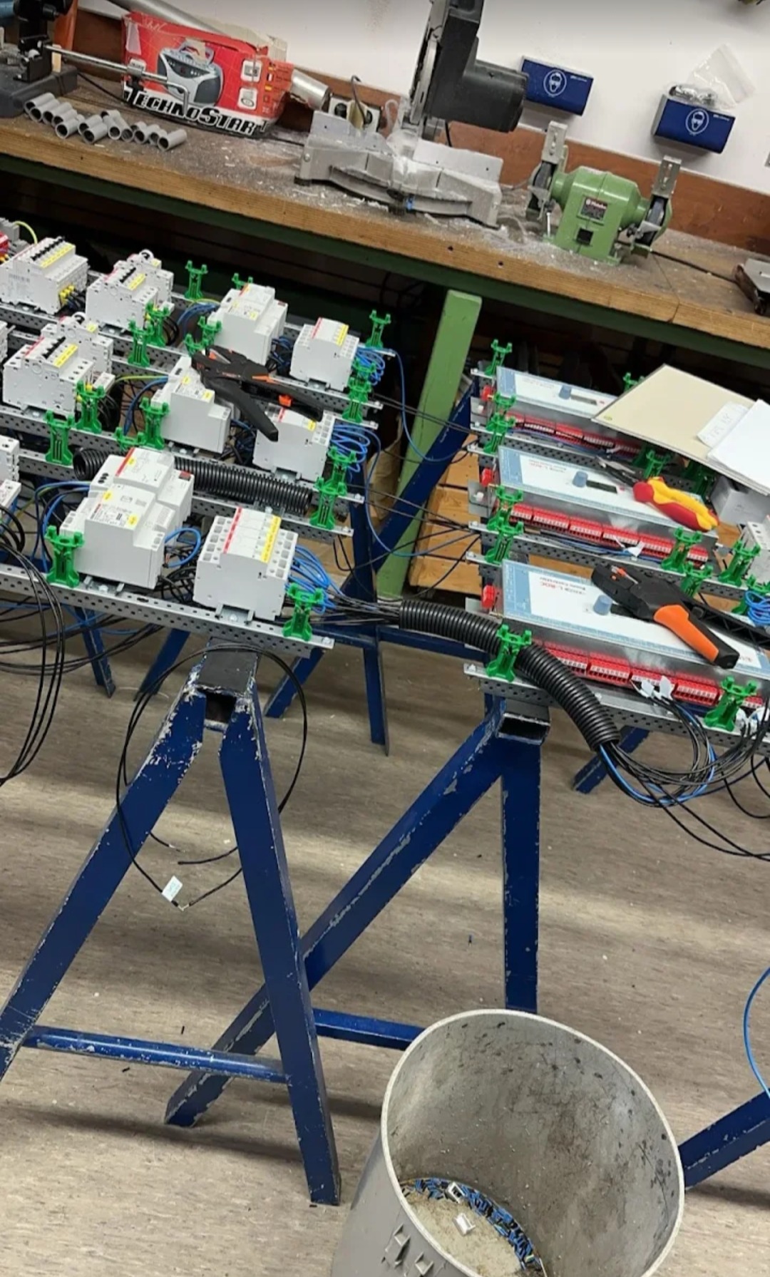 Rozdzielnice elektryczne na stojakach warsztatowych, w trakcie montażu. Widoczne narzędzia i przewody. Ujęcie z dołu, z widocznym koszem na odpady.