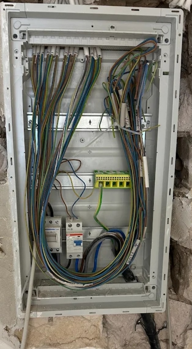 Rozdzielnica elektryczna w trakcie montażu: liczne przewody w różnych kolorach, zabezpieczenia i listwa zaciskowa w białej obudowie, surowy mur w tle.