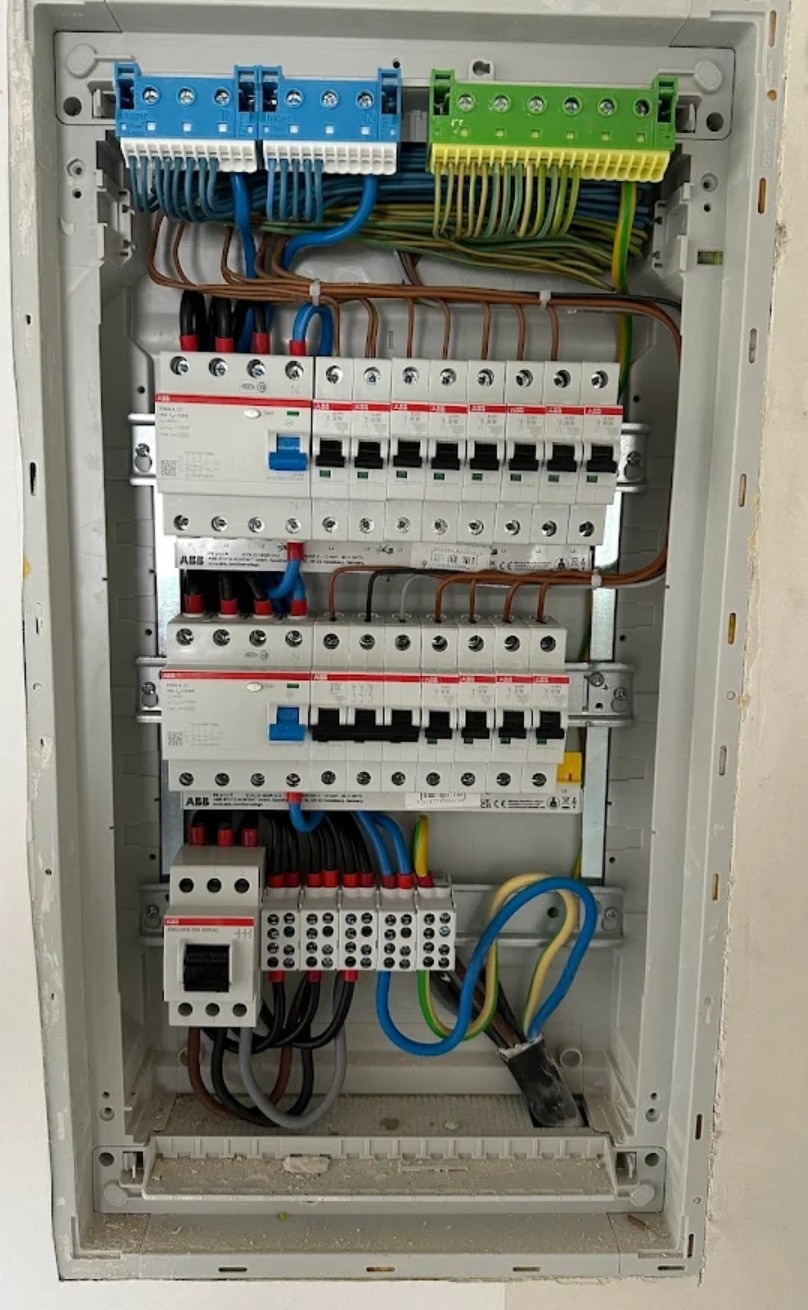 Rozdzielnica elektryczna z zainstalowanymi bezpiecznikami i przewodami w kolorach: niebieskim, brązowym, zielono-żółtym. Widoczne listwy zaciskowe i aparatura ABB.