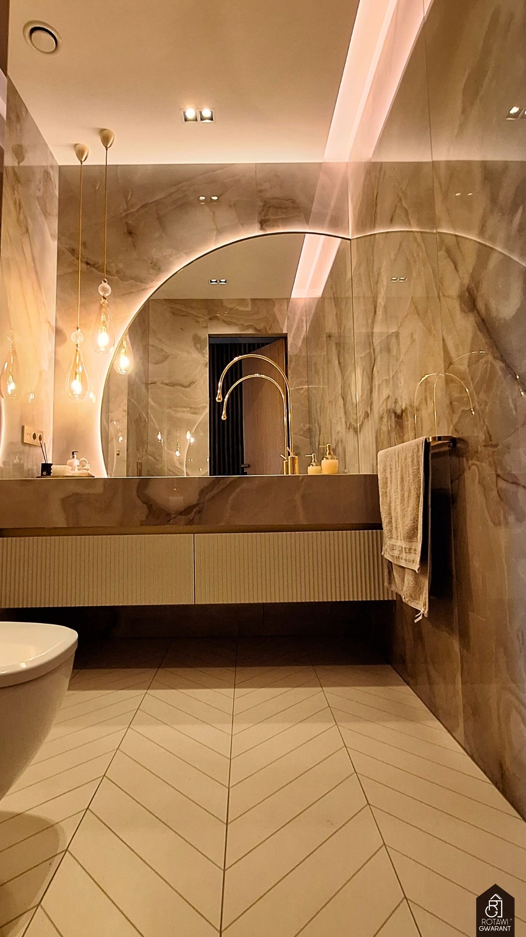 Elegancka łazienka z marmurowymi ścianami, okrągłym lustrem z podświetleniem LED, złotą armaturą i wzorzystą podłogą w jodełkę. Widoczny fragment toalety.