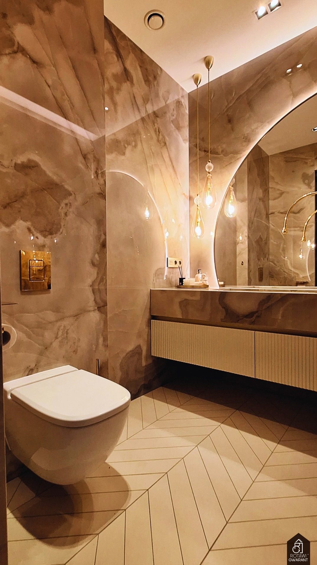 Elegancka łazienka z marmurowymi ścianami, podwieszaną toaletą i geometryczną podłogą. Złote akcenty: armatura, oświetlenie i rama lustra. Nowoczesny design.