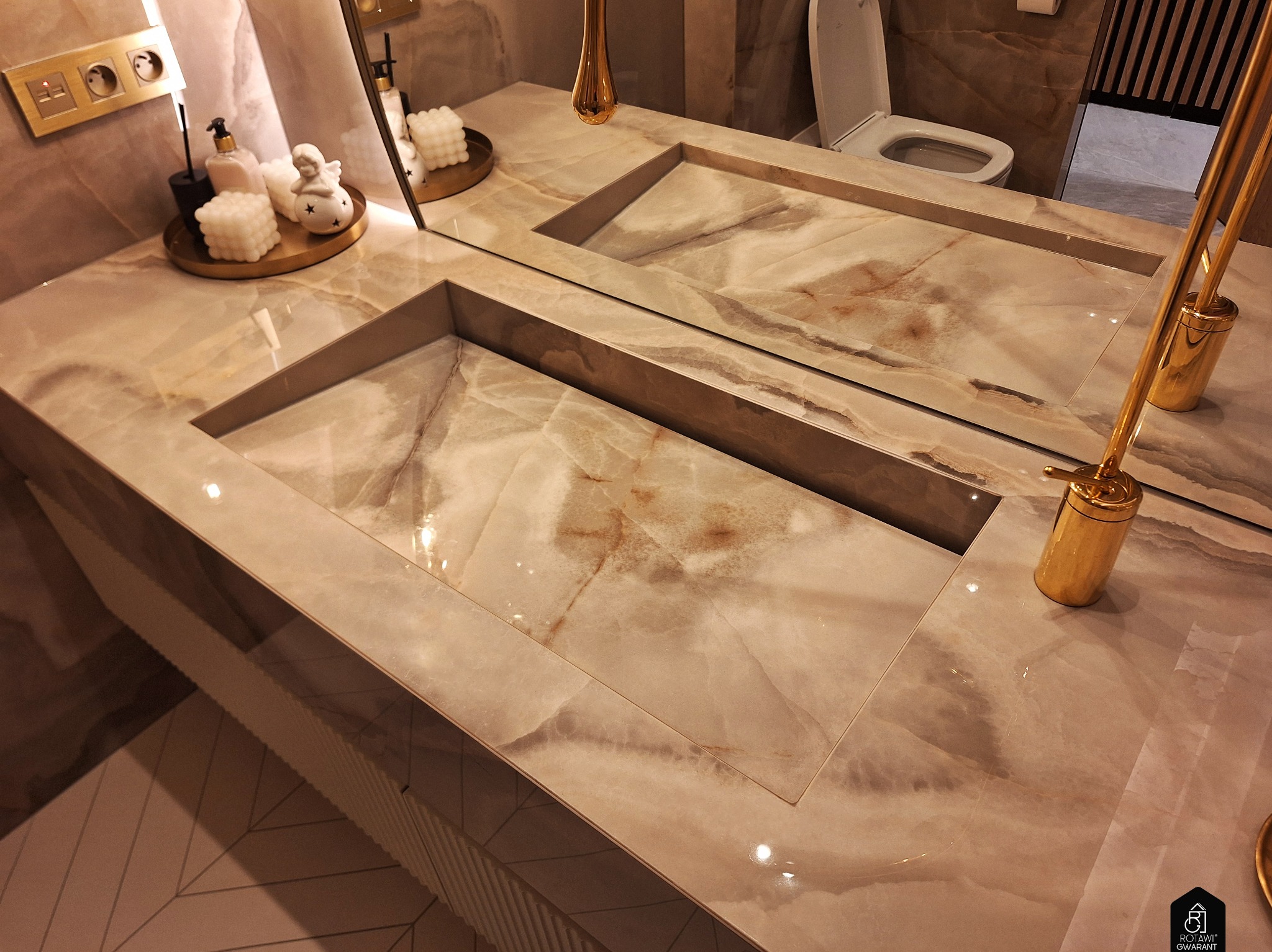Beżowa łazienka z umywalkami wpuszczonymi w marmurowy blat. W lustrze odbija się toaleta. Złote akcenty: dozownik, dekoracje. Nowoczesny design, ciepłe oświetlenie.