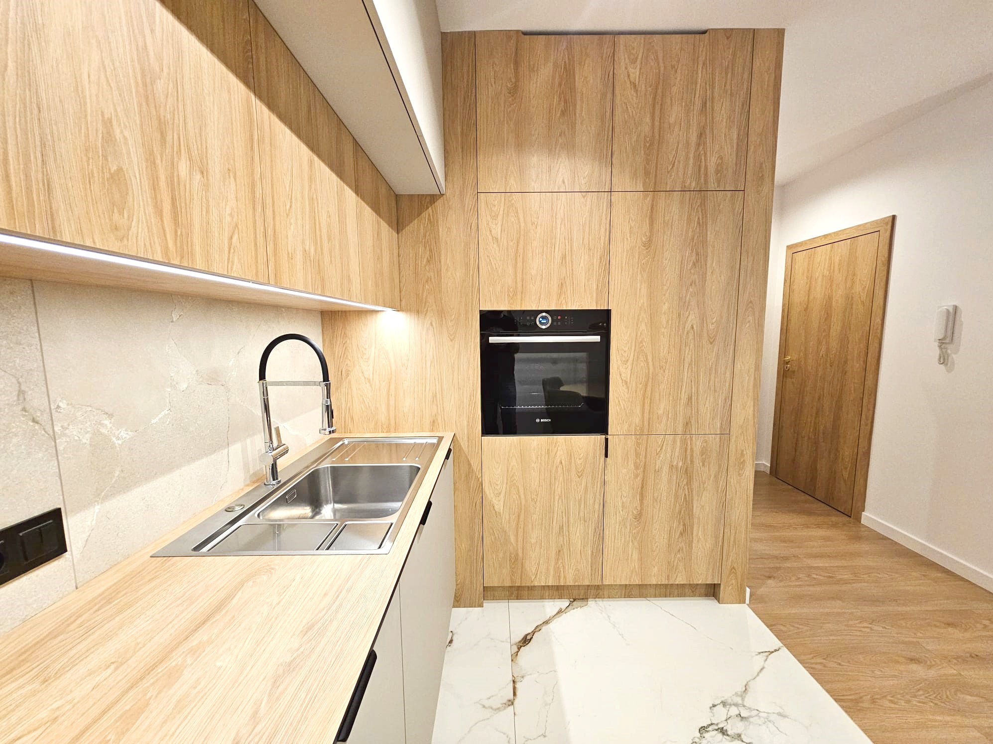 Nowoczesna kuchnia z drewnianymi frontami szafek, marmurową podłogą i oświetleniem LED. Ujęcie na zlew, baterię i zabudowany piekarnik. Widoczny fragment korytarza.