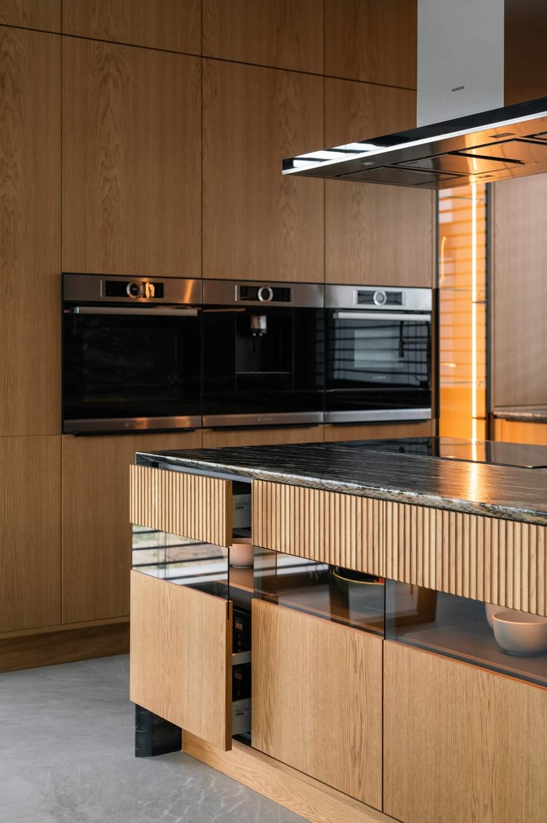 Drewniana zabudowa kuchenna z czarnym blatem i wbudowanymi sprzętami Siemens. Otwarta szuflada z oświetleniem. Nowoczesny design, ciepłe oświetlenie.