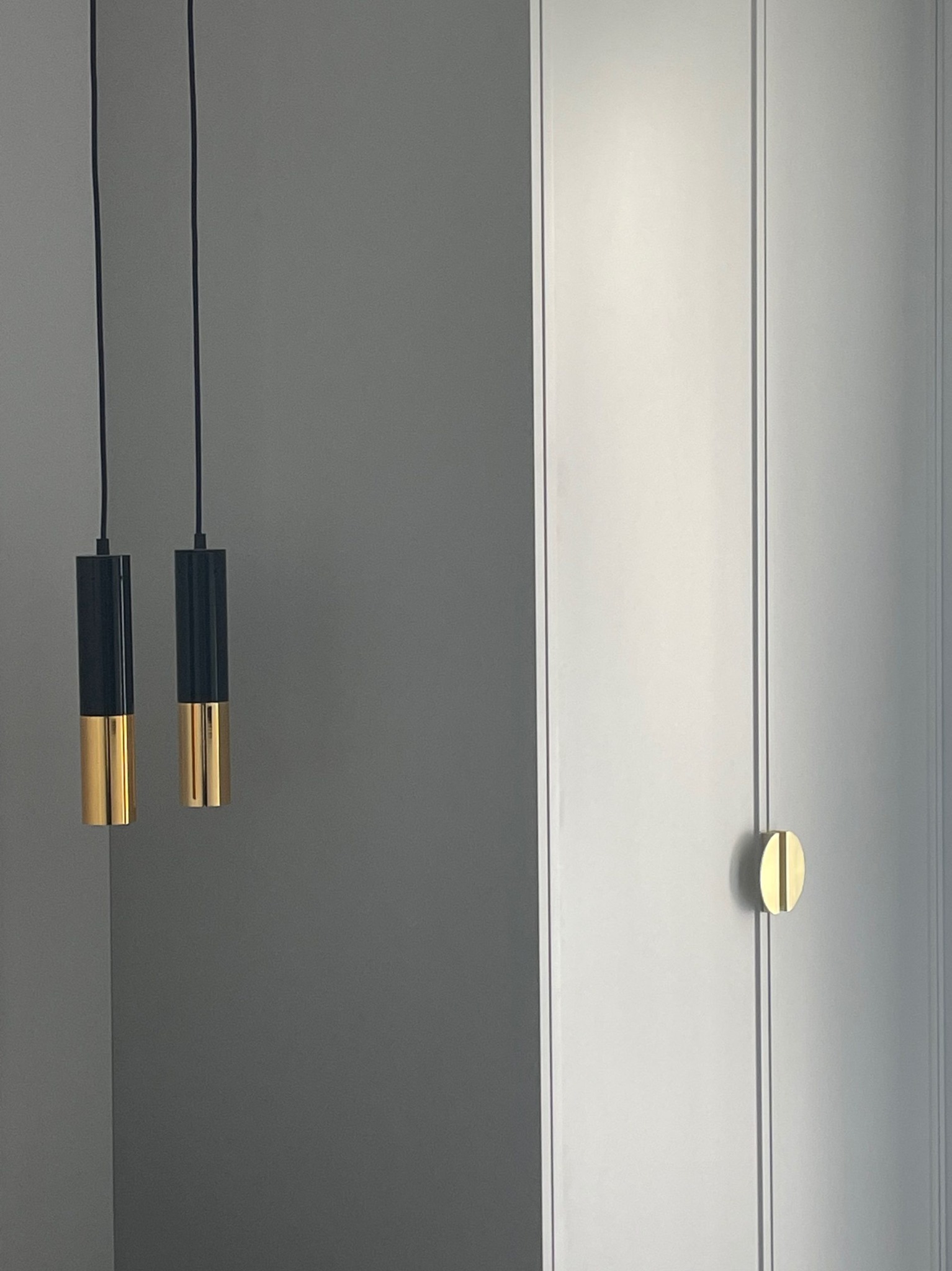 Nowoczesne wykończenie wnętrza: szara ściana, biała szafa z złotym uchwytem i dwie wiszące lampy z czarno-złotymi kloszami.