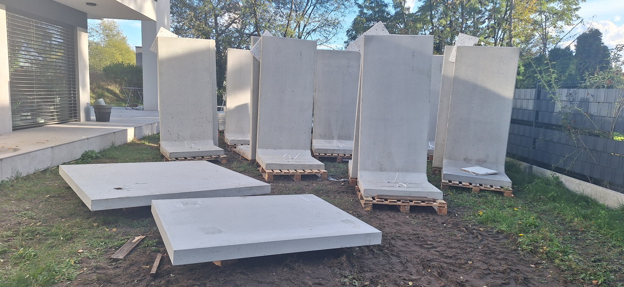 Betonowe prefabrykaty na paletach i płyty na ziemi, przygotowane do montażu na zewnątrz budynku. Widoczne elementy konstrukcyjne i otoczenie placu budowy.