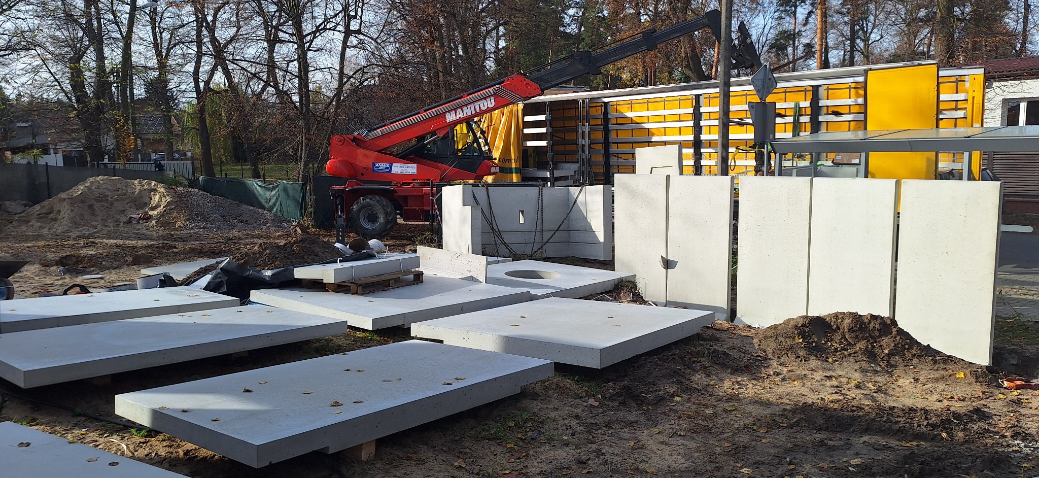 Montaż prefabrykowanych elementów betonowych na placu budowy z użyciem dźwigu Manitou. Widoczne płyty i ściany z betonu, przygotowane do instalacji.