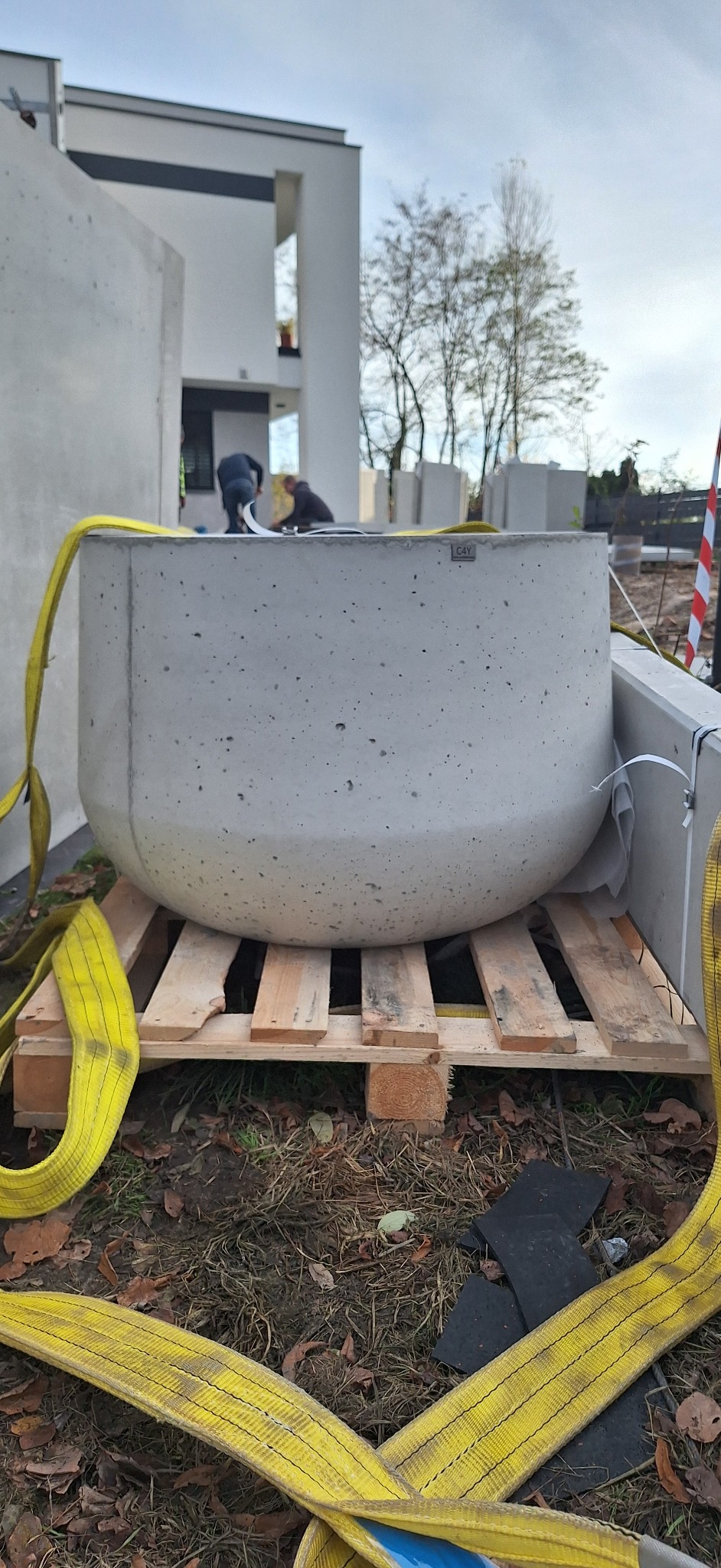 Betonowa donica w kształcie misy, umieszczona na palecie, z żółtymi pasami transportowymi. W tle budynek w trakcie budowy i pracujący robotnicy.